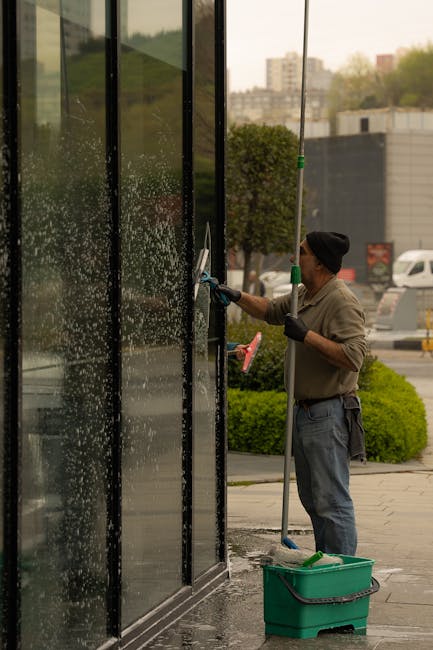 A professional worker using a high-pressure cleaning method to thoroughly clean large black glass panels on the exterior of a commercial building, with visible water spray and foam. The worker is dres