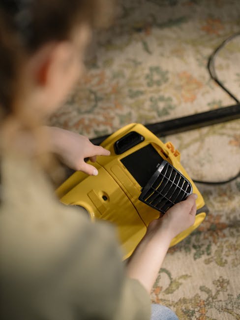 A person holding a yellow portable surface cleaning or sanitisation device, with black vent slats on the front, positioned over a textured, patterned carpet or rug with shades of beige, green, and ora