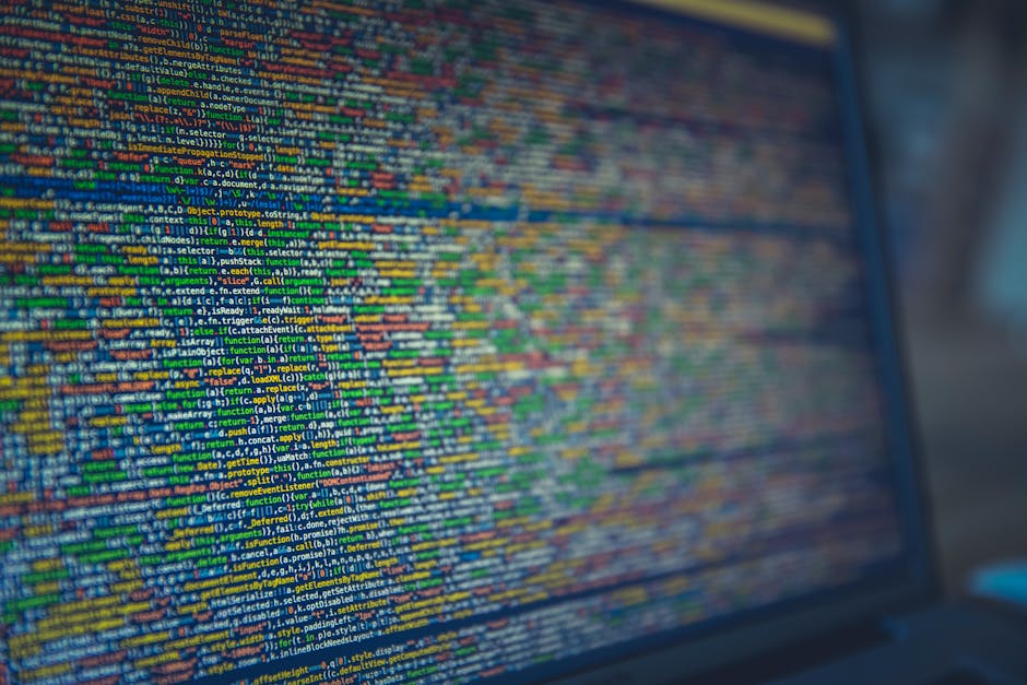 Close-up of a person's hands operating a laptop computer on a dark table, with the screen displaying lines of code in a code editor. The scene is well-lit, highlighting the keyboard and screen content